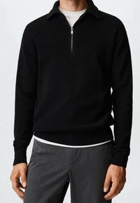 Pull noir côtelé avec un col zippé, manches longues et ourlet ajusté, porté sur une chemise blanche. Pantalon gris visible en dessous.