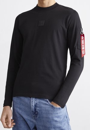 Mężczyzna ubrany w czarną koszulę z długim rękawem z czerwoną metką "Remove Before Flight" na kieszeni na lewym ramieniu oraz niebieskie dżinsy.
