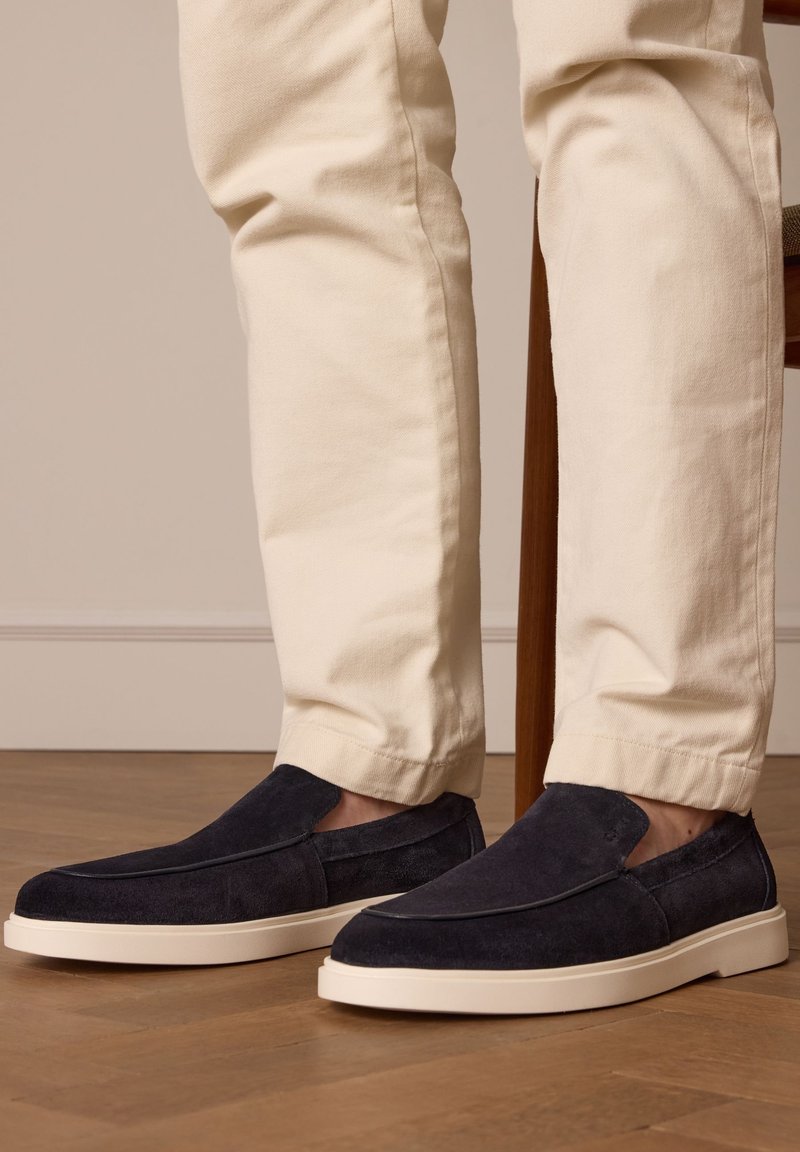 Homme portant un pantalon crème avec des chaussures en suède noir à enfiler, se tenant sur un sol en bois près d'une patte de chaise.