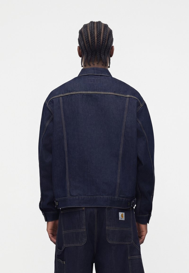 Person with braided hair wearing dark blue denim jacket and matching pants, viewed from the back against a plain background.