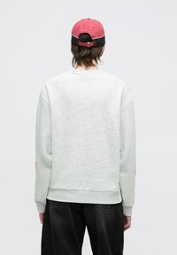 Gaiši pelēks džemperis ar apaļu kakla izgriezumu, nolocītām plecām, ribotām aprocēm un kontrastējošu sarkanu cepuri. Skats no muguras atklāj vienkāršu, tīru dizainu.