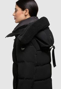 Geacă puffer neagră cu glugă pufoasă și guler interior lucios. Prezintă o patch cu logo rotund pe mânecă și bretele ajustabile.
