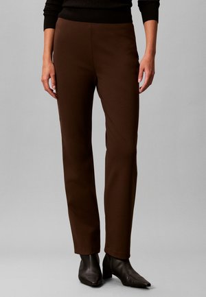 Personne portant un pantalon droit marron foncé et des bottines noires, debout devant un fond gris uni.