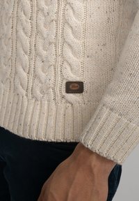 Gestrickter Creme-Pullover mit einem Zopfmuster und ribbeligem Saum, versehen mit einem braunen Lederetikett an der Seite. Die Textur ist dick und gemütlich.