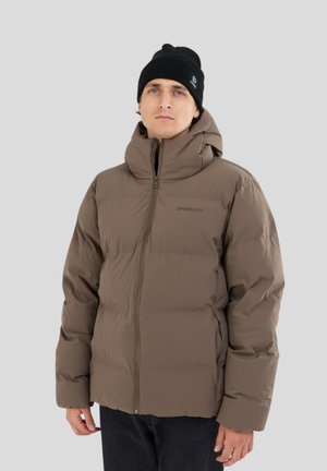 Jeune homme portant une veste matelassée marron avec un col haut et un bonnet noir, debout devant un fond gris clair uni.