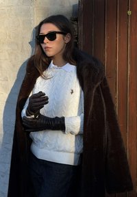 Woman wearing black sunglasses, black leather gloves, white cable knit sweater, dark fur coat draped over shoulders, standing by wooden door.