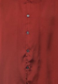 Camicia rossa con chiusura a bottoni, caratterizzata da una texture liscia, sei bottoni neri sul davanti e un design di colletto classico.