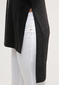 Maglietta nera a maniche lunghe con spacco laterale, abbinata a jeans bianchi. La maglietta presenta una consistenza morbida e una vestibilità rilassata.