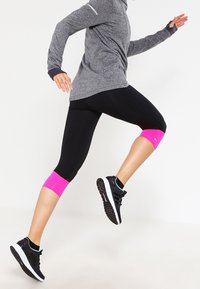 Osoba ubrana w szaro-skalny długi rękaw, czarne legginsy z różowymi mankietami i czarne buty do biegania, w trakcie biegu na jednolitym tle.