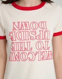 Κρεμ T-shirt με κόκκινο περιθώριο λαιμού και ανάποδο κείμενο "ΚΑΛΩΣ ΗΡΘΑΤΕ ΣΤΟ ΑΝΤΙΘΕΤΟ" σε έντονη, παιχνιδιάρικη γραμματοσειρά.