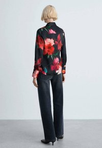 Blusa nera con grandi motivi floreali rossi, maniche lunghe e colletto, abbinata a pantaloni in denim scuri a vita alta.