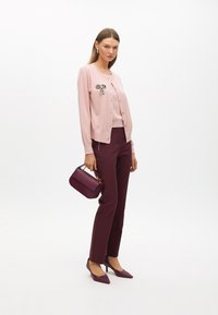 Ljusrosa cardigan med en rosettbrosch, kombinerad med vinröda byxor och matchande spetsiga klackar. Inkluderar en liten vinröd handväska.