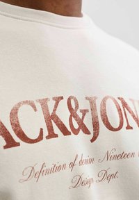 Sweatshirt en coton beige clair avec un grand texte rouge indiquant "BLACK&JONES" et un texte plus petit en dessous. Col rond côtelé.