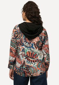 Färgglad, mönstrad hoodie med en svart huva, som har livfulla blommönster och abstrakta designer i orange, grön och blått på en vit bakgrund.