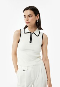 Ärmlös vit ribbad topp med krage, svart kant och mittdragkedja. Bärs med ljusfärgade byxor, vilket ger en avslappnad look.