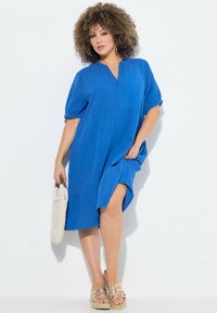 Abito camicia blu con maniche corte, apertura frontale con bottoni e tasche; abbinato a sandali con plateau dorati e una borsa di paglia beige.