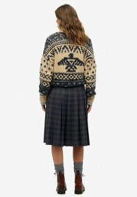 Pull en tricot avec une base crème et des motifs bleu foncé, comprenant un emblème central ; associé à une jupe plissée à carreaux et des bottes marron.