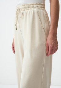 Beige joggers met een elastische tailleband en trekkoord. Gladde stof met zijzakken en een relaxte pasvorm, weergegeven vanaf de zijkant.