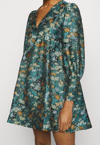 Kurzes Kleid aus türkisfarbener Stoff mit einem lebhaften Blumenmuster in den Farben Orange, Blau und Grün; V-Ausschnitt und geraffte Ärmel.