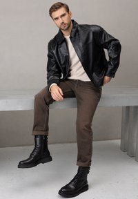 Czarna skórzana kurtka, beżowy sweter, brązowe dżinsy oraz czarne sznurowane buty z zamkami. Gładka tekstura, dopasowany krój i podwinięte mankiety.