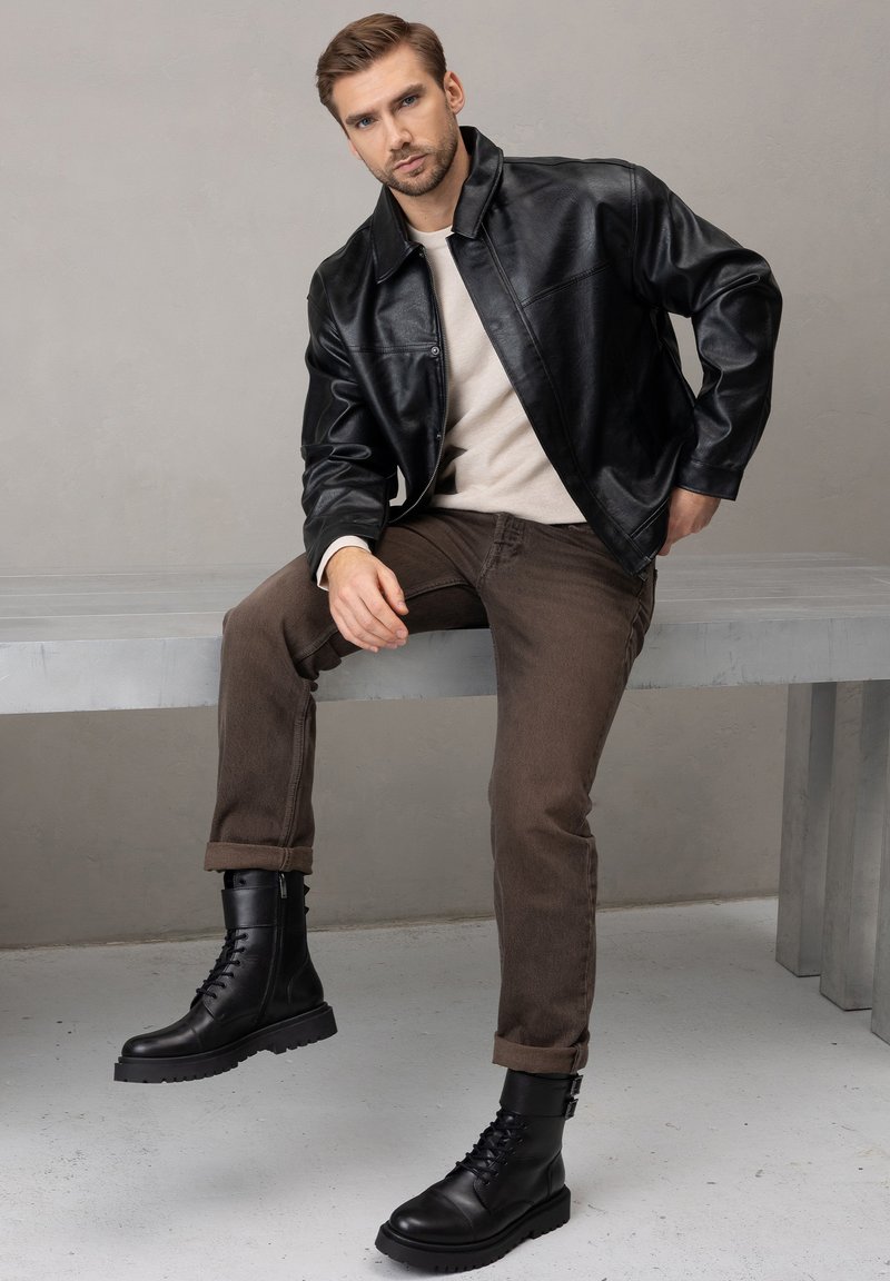Czarna skórzana kurtka, beżowy sweter, brązowe dżinsy oraz czarne sznurowane buty z zamkami. Gładka tekstura, dopasowany krój i podwinięte mankiety.