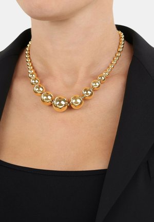 Collana in oro con perline a goccia lucide e sfaccettate, indossata su una maglietta nera e un completo.