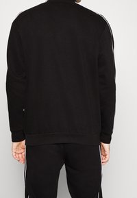 Veste de sport noire avec col montant et bandes blanches sur les côtés, dotée de poignets et d'un ourlet côtelés, confectionnée en tissu doux et extensible.