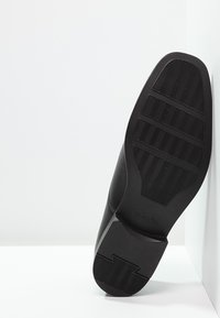 Chaussure en cuir noir avec une semelle en caoutchouc texturée présentant un motif en chevrons. L'arrière de la chaussure est incliné sur un fond blanc.