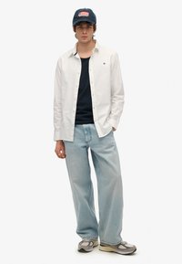 Camisa blanca de botones sobre una camiseta sin mangas azul marino, combinada con jeans de pierna ancha azul claro y zapatillas grises, rematada con una gorra azul marino con un diseño de bandera.