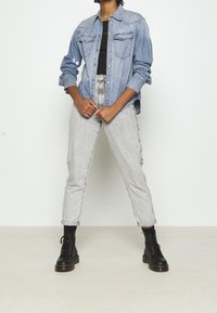 Jasnoniebieska koszula dżinsowa z dwoma przednimi kieszeniami, zestawiona z jasnoszarymi spodniami o wysokim stanie. Model nosi czarne buty na sznurówki.