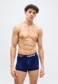  Νεαρός άνδρας με σγουρά μαλλιά, φορώντας μπλε μποξεράκια Nike, στέκεται μπροστά σε απλό ανοιχτό φόντο, κοιτάζοντας προς τα εμπρός.