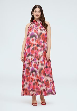 Femme portant une robe maxi sans manches à col halter avec des motifs floraux rouges, roses et violets et des talons ouverts rouges sur un fond uni.