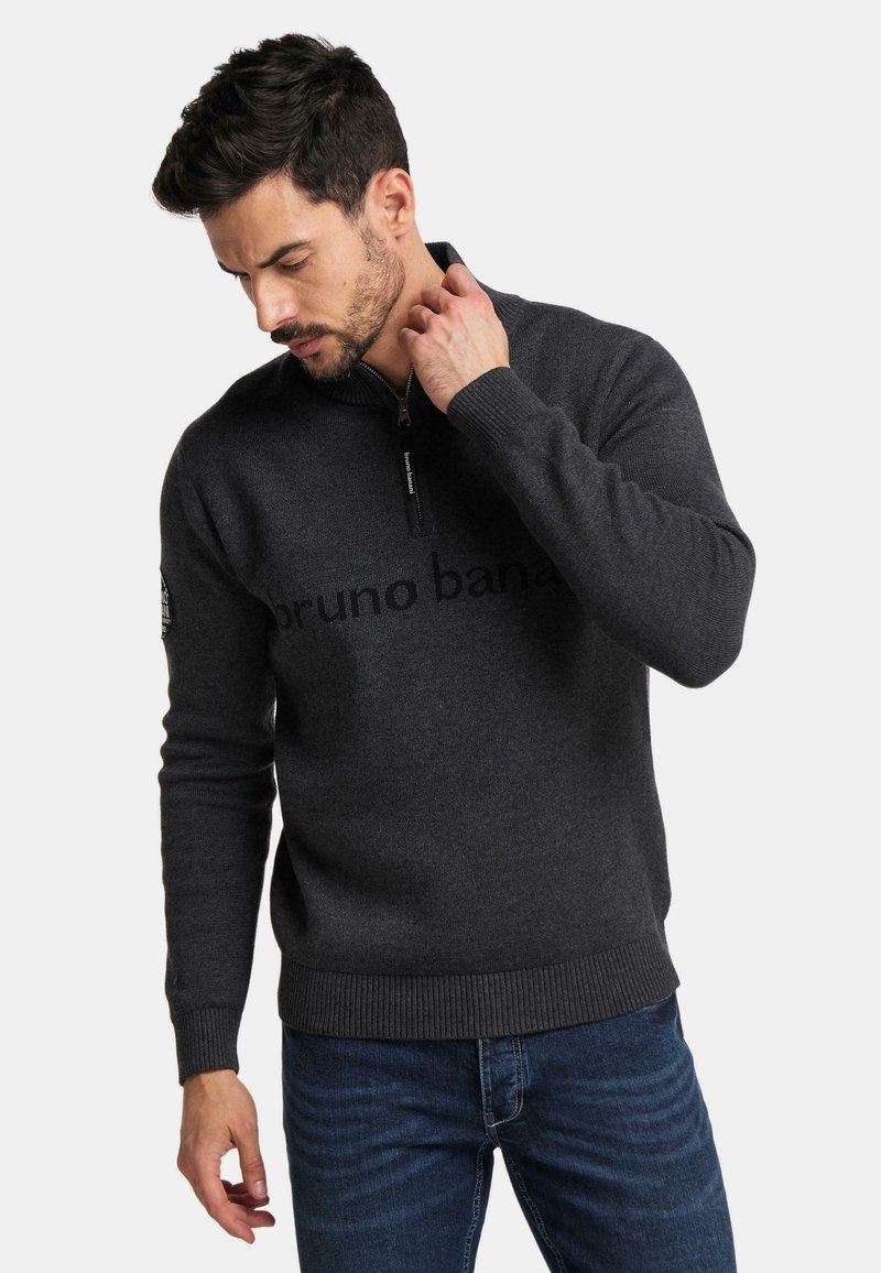 Dunkelgrauer Reißverschluss-Pullover mit hohem Kragen, der mit einem dezent gedruckten Logo versehen ist. Ribbelbündchen und -saum sorgen für Struktur. Kombiniert mit Jeans.