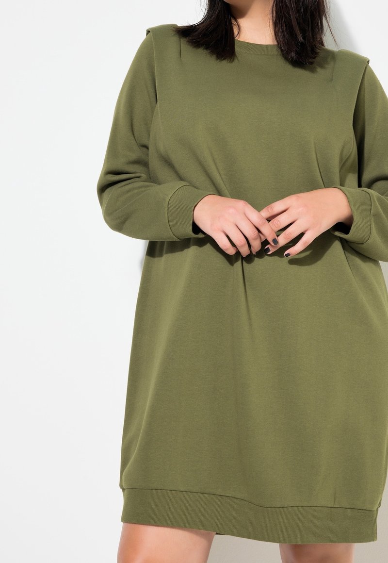 Femme portant une robe-pull à manches longues vert olive avec des poignets côtelés, debout devant un fond blanc uni.