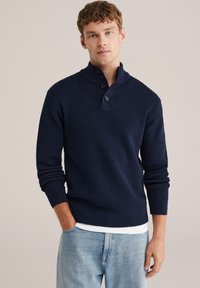 Mörkblå tröja i strukturstickad tyg med hög krage, tre knappar vid halsen och ribbade ärmmuddar. Bärs över en vit skjorta.