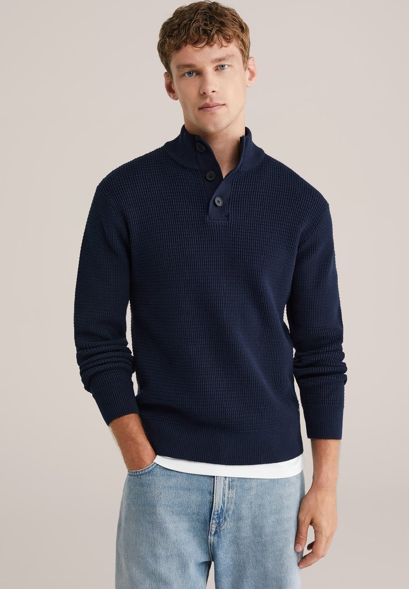 Mörkblå tröja i strukturstickad tyg med hög krage, tre knappar vid halsen och ribbade ärmmuddar. Bärs över en vit skjorta.
