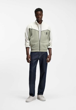 Man wearing a color-block zip-up jacket, dark jeans, and light sneakers standing against a plain white background.