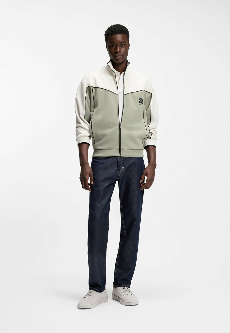 Homme portant une veste zippée à blocs de couleurs, un jean foncé et des baskets claires, debout devant un fond blanc uni.