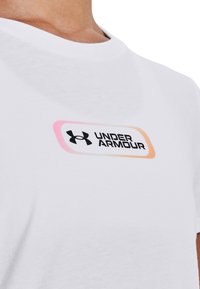 Vit bomull t-shirt med en rektangulär gradientlogotyp där "UNDER ARMOUR" är i svart med nyanser av rosa och orange som accenter.