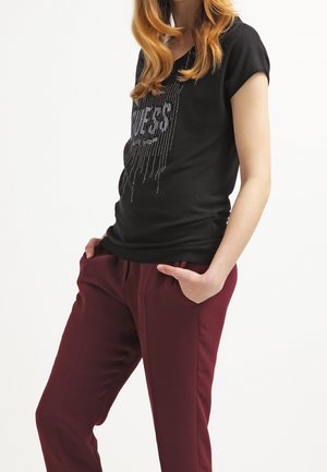 Musta T-paita, jossa on tekstuurilla varustettu graafinen kuvasto, jossa on sana "Jeans" hopeisista kimalteista, yhdistettynä viininpunaisiin kapeneviin housuihin.