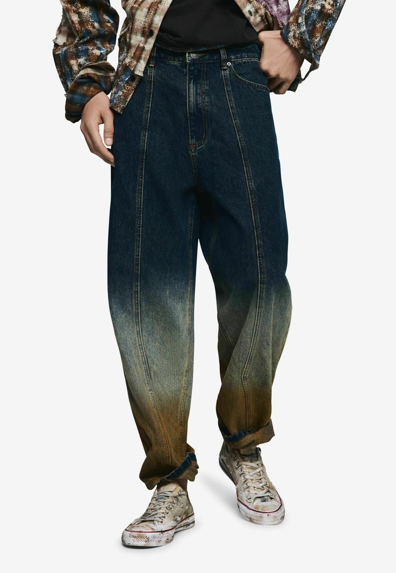 Farmernadrág, amely sötétkéktől világosig fakul lefelé, oldalsó varrási részletekkel és kényelmes szabással. Kopott tornacipővel viselve.