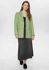 Grön texturerad blazer med en strukturerad design, kombinerad med en svart kjol. Outfitten har subtila sömnadsdetaljer och svarta skor.
