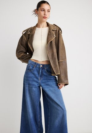 Braune Leder-Bikerjacke mit breiten Revers, kombiniert mit einem enganliegenden cremefarbenen Rippentop und locker sitzenden blauen Jeans mit hohem Bund und weitem Bein.