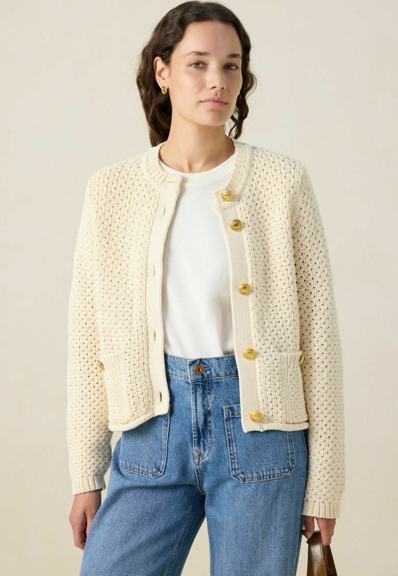 Cardigan en tricot crème avec un motif texturé, col rond et boutons dorés. Associé à un jean bleu taille haute et un sac à main.