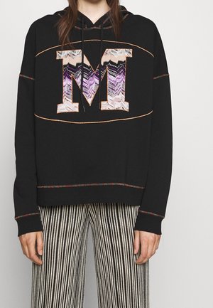 Sweat à capuche noir avec un grand "M" texturé présentant des motifs en zigzag en violet et beige. Des coutures vives et une coupe décontractée définissent le design.