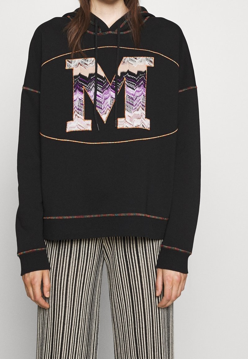 Schwarzer Hoodie mit einem großen, strukturierten "M", das Zickzackmuster in Lila und Beige aufweist. Helle Nähte und eine lockere Passform zeichnen das Design aus.