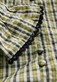 Tissu à carreaux vert, jaune et marron avec boutons recouverts du même tissu et bordure noire nouée sur la zone du poignet froncée.