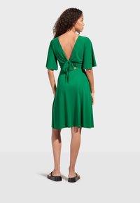Femme aux cheveux bouclés portant une robe verte à manches courtes avec un dos ouvert noué en un nœud, debout en sandales noires sur un fond uni.