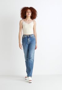 Lichtbeige top met kantafwerking, gecombineerd met hooggzette blauwe spijkerbroeken. Het model draagt witte plateau-sandalen en staat voor een witte achtergrond.