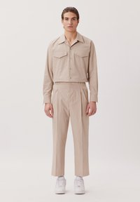 Chemise beige à manches longues avec deux poches poitrine, assortie à un pantalon taille haute beige et des baskets blanches. Tissu lisse, coupe ajustée.